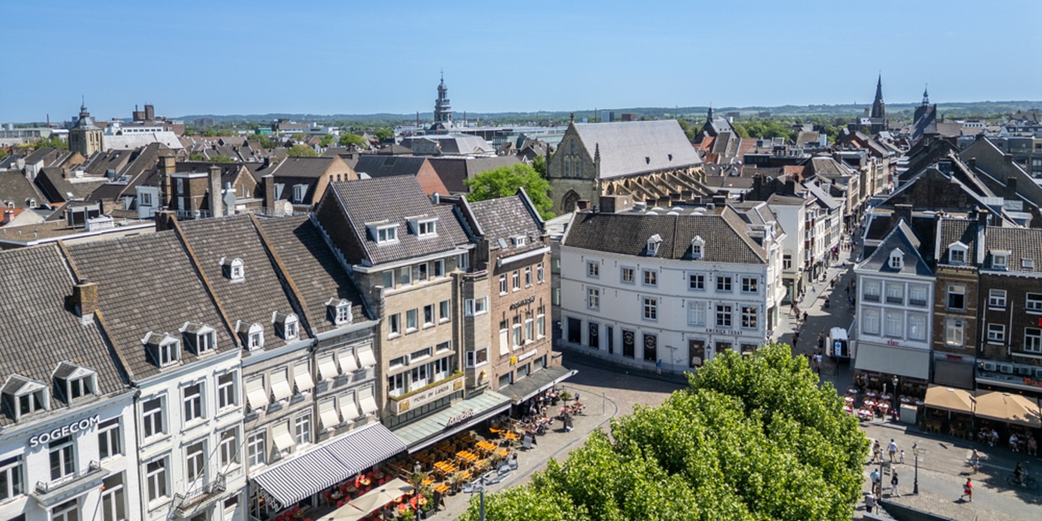 Het Vrijthof in Maastricht bij zonsondergang, met op de achtergrond de imposante Sint-Servaasbasiliek en de roodverlichte Sint-Janskerk, omringd door historische gebouwen en een muziekkiosk op het plein.