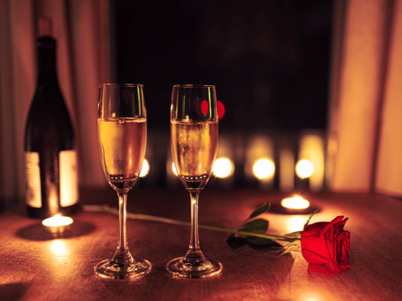 Two filled champagne glasses on a wooden table with a red rose and burning candles, in the background a bottle of wine and soft ambient lighting for a romantic setting.